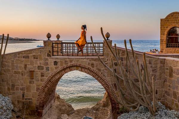 Frau in einem orangefarbenen Kleid auf einem Steinbogen am Meer