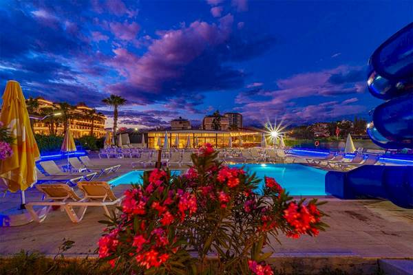 Abendansicht des Poolbereichs im Club Wasa Holiday Village mit beleuchteten Liegestühlen und Blumen.