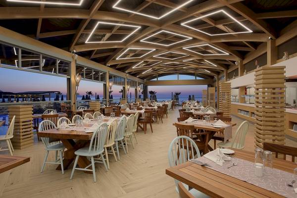 Innenansicht des Restaurants im Arina Beach Resort mit Holztischen und Stühlen, Glaswänden und einer Meeresaussicht.