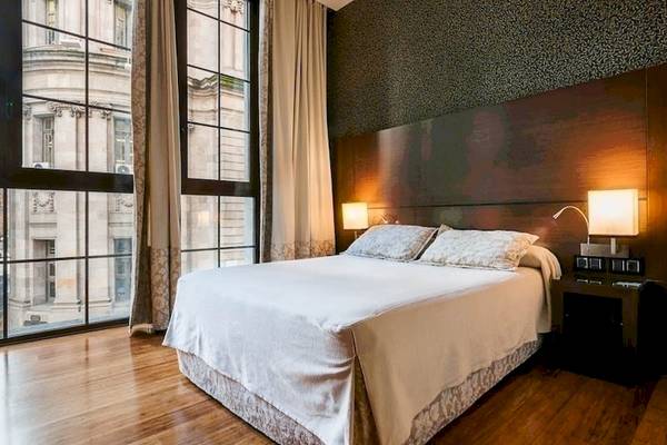 Schlafzimmer im Colonial Barcelona Hotel mit großem Bett, Nachttischen und Fenstern mit Blick auf ein Gebäude.