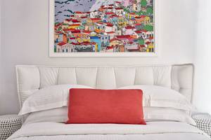 Bett mit weißen Kissen und roter Kopfstütze im Schlafzimmer mit einem Wandbild des Myconian Ambassador Hotels.