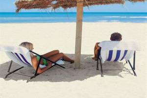 Zwei Personen sitzen unter einem Strohhut auf dem Strand, einer liest ein Buch.