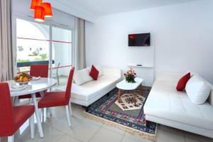 Wohnzimmer im Djerba Holiday Beach Hotel mit weißen Möbeln, rotem Stuhl, Tisch und Fernseher an der Wand.