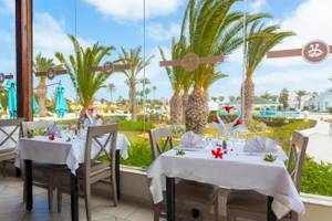 Innenansicht des Restaurants des Djerba Holiday Beach Hotels mit Tischen und Stühlen, Palmen und einem Swimmingpool im Hintergrund.