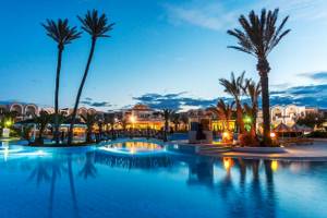 Ein Swimmingpool des Djerba Holiday Beach Hotels bei Nacht, umgeben von Palmen und Gebäuden.