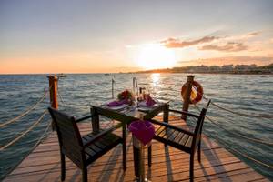 Ein Tisch auf einer Holzplattform am Wasser bei Sonnenuntergang, mit Flaschen und Blumenarrangement.