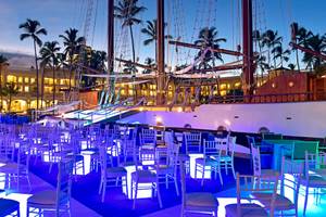 Abendliche Feier auf der Terrasse des JOIA Bávaro by Iberostar Hotel mit blau beleuchteten Stühlen und einem Schiff im Hintergrund.