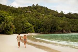 Ein Paar geht am Strand des Constance Ephelia Resorts entlang, umgeben von grüner Vegetation und kristallklarem Wasser.