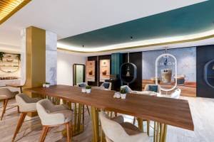 Innenbereich des Nirvana Cosmopolitan Hotels mit einem langen Holztisch, Sesseln, Pflanzen und einer Wand mit einer Wanddekoration.