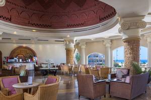 Innenbereich des Utopia Beach Club Hotels mit runden Tischen und Stühlen, einer Bar und großen Fenstern.