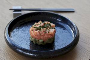 Lachs-Avocado-Tartare auf einem schwarzen Teller, auf einem Holztisch serviert, mit Essstäbchen.