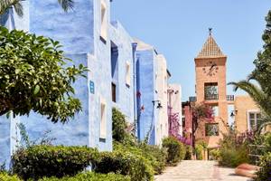 Haupteingang des Candia Park Village Hotels mit blauen Gebäuden und einer Uhrturm im Hintergrund.
