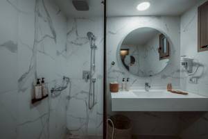 Ein Badezimmer im Eliros Mare Hotel mit einer weißen Marmorwand, einer Glasdusche, einem Spiegel und einem Waschbecken.