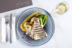 Gegrillter Fisch auf Teller mit Gemüse und Weingläsern auf weißem Tisch