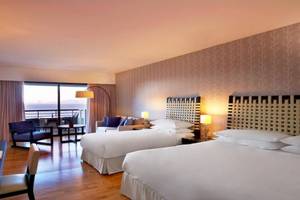 Doppelzimmer mit Meerblick im Sheraton Rhodes Resort mit zwei Betten, Schreibtisch, Sesseln, Couch, Fenstern und Balkon.
