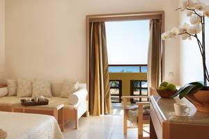 Grecotel Meli Palace Zimmer mit Sofa und Meerblick, das Sofa ist mit Kissen ausgestattet und es gibt einen Tisch mit einer Tasse, einen Nachttisch und eine Vase mit weißen Orchideen.