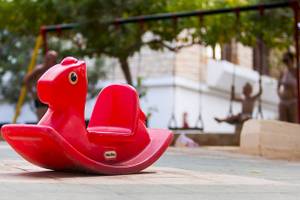 Ein rotes Schaukelpferd im Freien, im Hintergrund sind Kinder auf Schaukeln zu sehen.