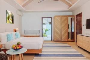 Schlafzimmer im Nova Maldives Hotel mit weißem Bett, Holzmöbeln und blau-weiß gestreiftem Teppich.