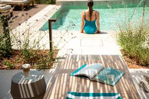 Eine Frau in einem blauen Badeanzug sitzt an einem Pool am Rand und liest ein Buch.