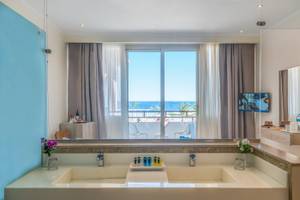 Badezimmer-Ansicht im Three Corners Equinox Beach Resort mit großem Fenster, das auf den Ozean und eine Terrasse mit Stühlen hinausblickt.