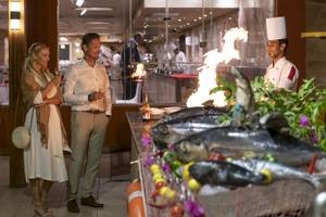 Zwei Gäste im Three Corners Happy Life Beach Resort halten Gläser mit Wein, während ein Koch ein Gericht anzündet.