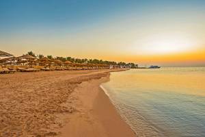 Sonnenuntergang am Strand des SUNRISE Royal Makadi Resort mit leeren Sonnenliegen und einem Boot im Wasser.