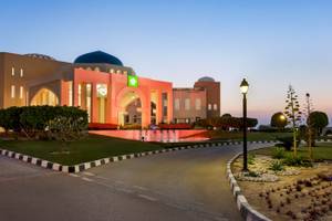 Aussenansicht des Wyndham Garden Salalah Mirbat Hotels bei Sonnenuntergang mit Brunnen und Lampen.