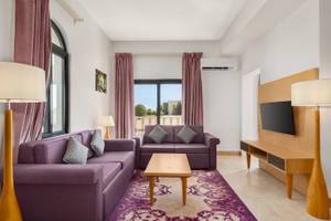 Wohnzimmer im Wyndham Garden Salalah Mirbat mit zwei lila Sofas, einem Tisch, Lampen und einem Fernseher.