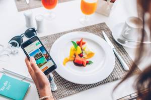 Eine Frau hält ein Smartphone mit der Website von Iberostar Waves Alcudia Park vor einem Frühstückstisch.