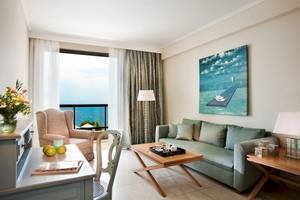 Wohnzimmer im Ikos Oceania Hotel mit Sofa, Tisch, Blumenvase und Meerblick.