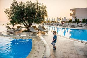 Ein Kind steht am Pool im Ikos Oceania Hotel neben einem Baum und vielen Liegen.