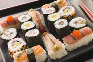 Eine Auswahl verschiedener Sushi-Rollen auf einem schwarzen Teller, mit Stäbchen daneben.