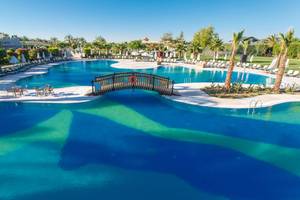 Pool des Sherwood Exclusive Lara Hotels mit Sonnenliegen, Schirmen und einer Brücke.