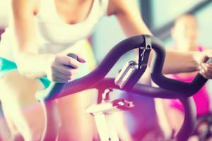 Frau im Fitnessstudio auf einem Fahrrad, während eine weitere Person im Hintergrund ebenfalls fährt.