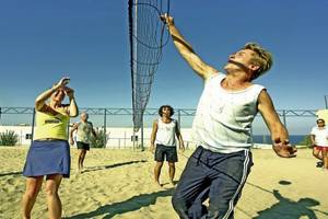 Eine Gruppe von Menschen spielt Volleyball auf einem Sandplatz unter einem blauen Himmel.