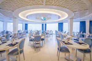 Innenbereich des Adams Beach Hotels, Restaurant mit runden Tischen und Stühlen, zentrale Beleuchtung und blaue Vorhänge.