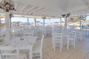 Innenbereich des Adams Beach Hotel Restaurants mit weißen Tischen und Stühlen und Blick auf den Strand.