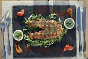 Gegrillter Fisch auf schwarzem Teller mit Rucola und Zitronenscheiben.