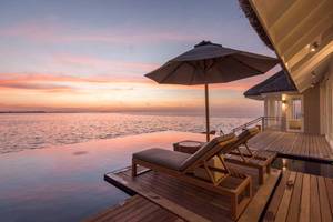 Terrasse des LUX South Ari Atoll Hotels mit zwei Liegen und einem Sonnenschirm am Meer bei Sonnenuntergang.