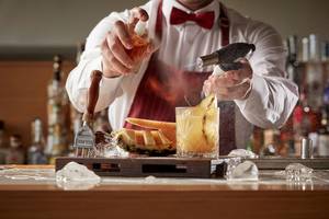 Barkeeper beim Avra Imperial Hotel bei der Zubereitung eines Cocktails mit einer Pinzette, einer Flasche und einer Fackel.