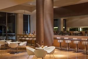 Innenansicht der Bar im Avra Imperial Hotel mit Holzboden, weißen Stühlen und einer Theke mit vielen Flaschen.