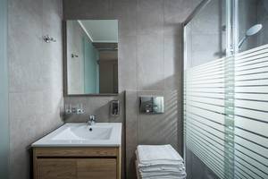 Ein Badezimmer im Odyssia Beach Hotel mit grauen Fliesen, einem Waschbecken, einem Spiegel und einer Glasdusche.