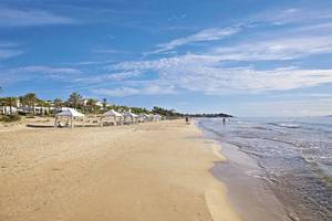 Strand des Grecotel La Riviera & Aqua Park mit weißen Sonnenschirmen und Hotels im Hintergrund.