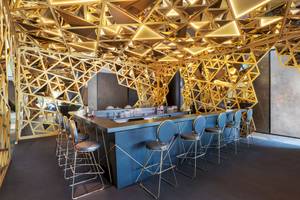 Bar im W Dubai - The Palm Hotel mit goldenen geometrischen Strukturen an der Decke und blauen Barhockern.
