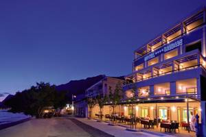 Das Saudade Hotel in der Abenddämmerung, mit beleuchteten Fenstern und einer Terrasse mit Tischen und Stühlen, die zum Strand führen.