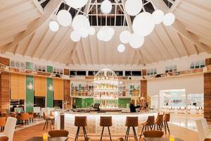 Lobby-Bar im H10 Playa Esmeralda Hotel mit einer Person hinter der Theke, umgeben von Holztischen und Stühlen.