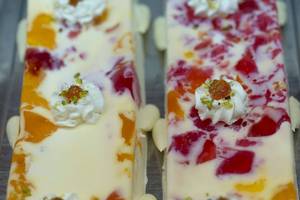 Nahaufnahme von zwei weißen Cremedesserts mit roten und orangefarbenen Fruchtstücken, dekoriert mit Pistazien und Schlagsahne.