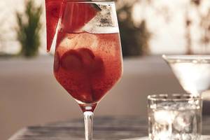 Ein Glas mit einem roten Cocktail und einem Stück Wassermelone auf einem Tisch mit weiteren Gläsern.