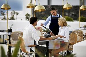 Zwei Gäste sitzen an einem Tisch im Freien des Santo Pure Oia Suites & Villas, während eine Kellnerin ihnen das Menü serviert.