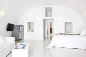 Innenansicht eines Schlafzimmers im Katikies Chromata Santorini Hotel mit weißem Bett, Couch, Tisch und Fernseher.